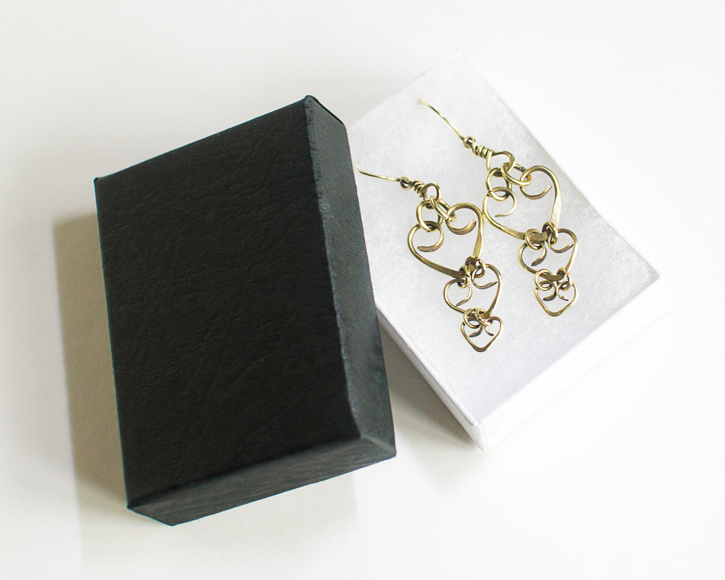 Brass heart-shaped earrings on a white card next to a black jewelry box on a white background