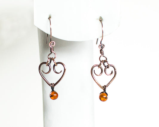 Heart-shaped copper earrings with amber beads on a white stand.